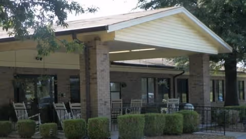 Exterior view of a covered patio area at Ripley Healthcare & Rehabilitation Center with brick pillars, outdoor chairs, tables, and neatly trimmed bushes in front. Trees provide shade around the building.