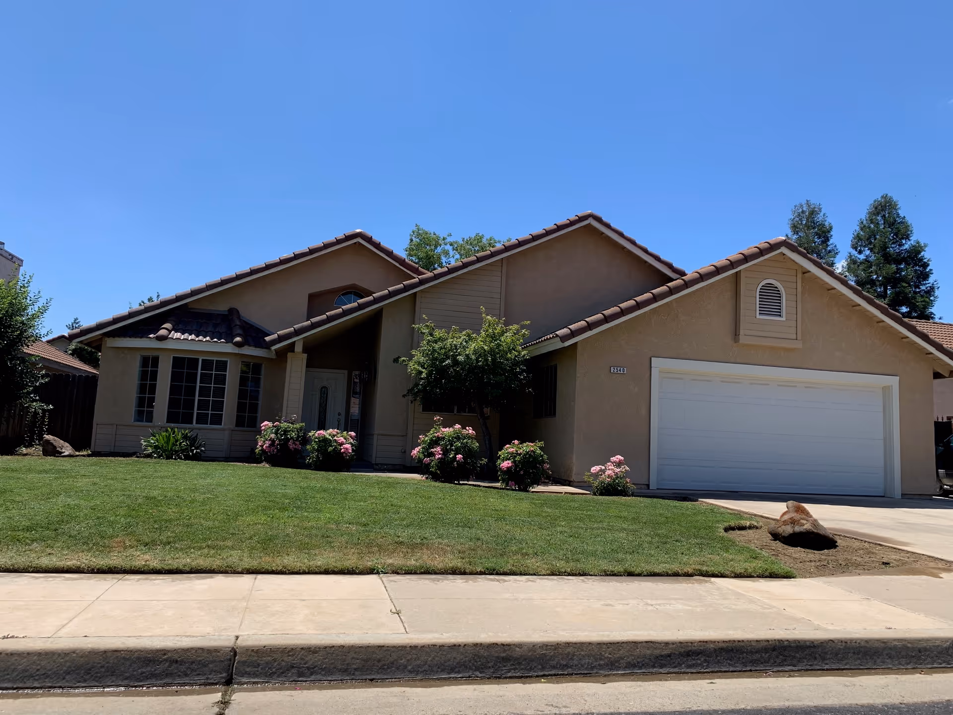 Single-story residential house with beige exterior walls and a brown tiled roof under a clear blue sky. The house features a large front lawn with green grass and several bushes with pink flowers near the entrance. There is a white garage door on the right side and a concrete driveway leading to it.