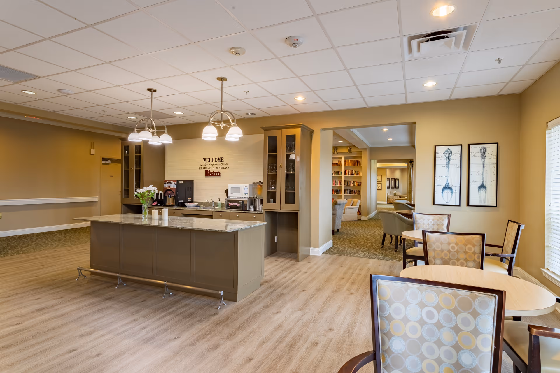 Open senior living common area with a central kitchen island, pendant lights, and tables and chairs next to a lounge and bookcase area.