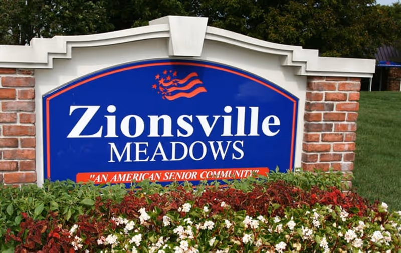 A large blue sign with white and red text reading 'Zionsville Meadows' and 'An American Senior Community' surrounded by a brick frame and greenery with flowers in front.