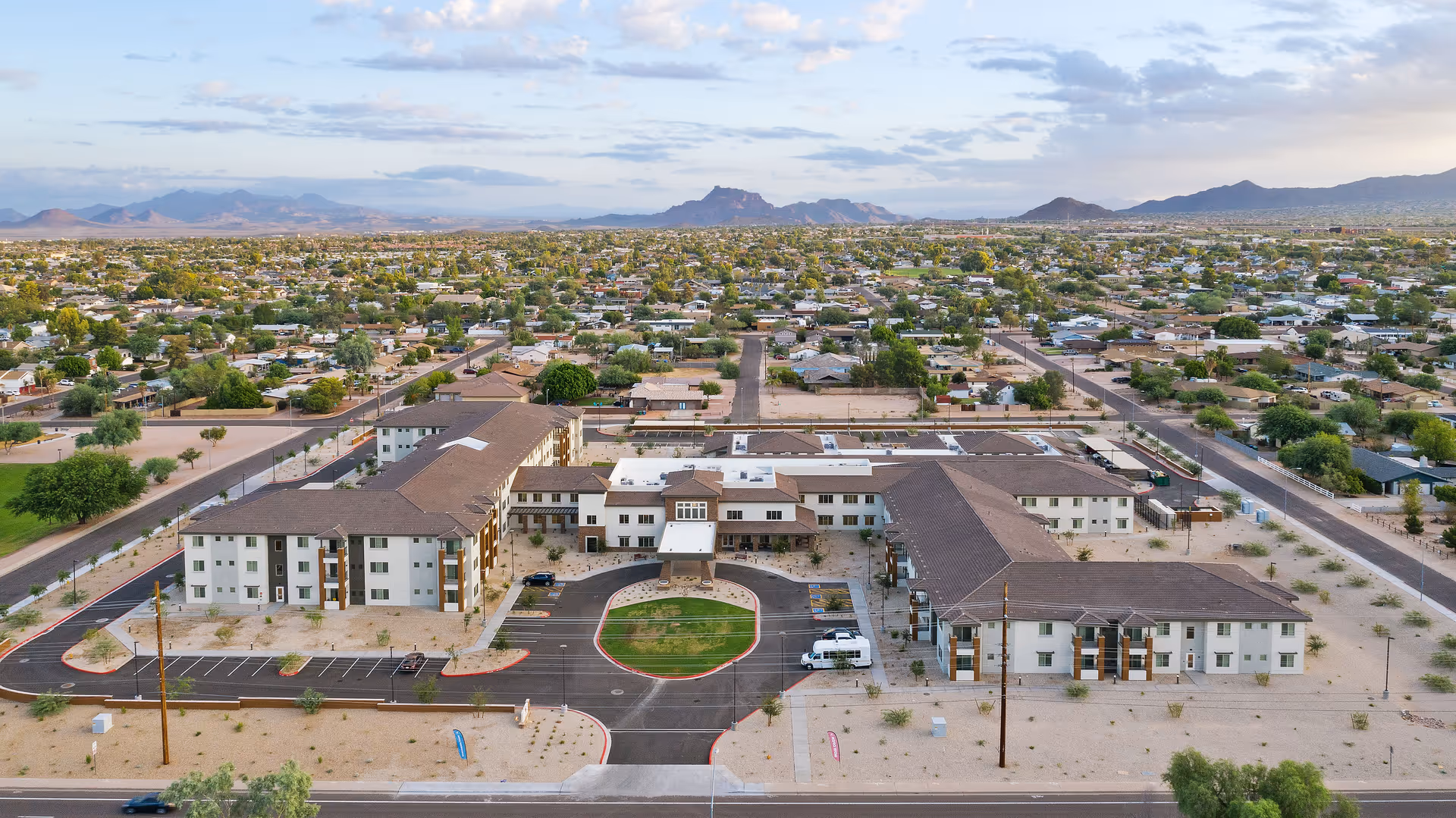 Aerial view of the Acoya Mesa senior living campus — a U-shaped, three-story building with a central circular drive, parking, desert landscaping, and a suburban neighborhood with mountains beyond.