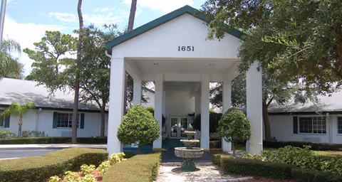 Front entrance of a senior living building with a white portico numbered 1651, a fountain, and landscaped shrubs.