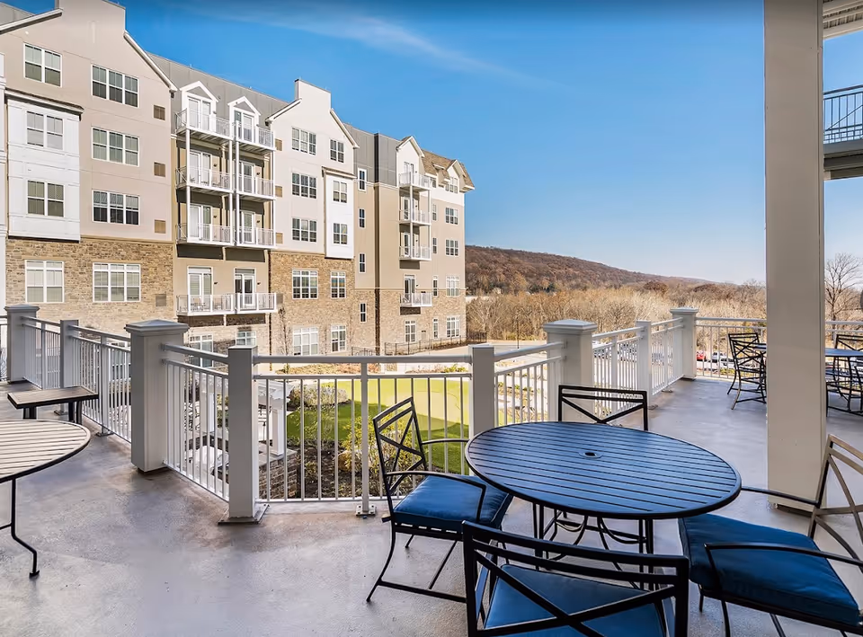 Outdoor balcony area with round tables and chairs with blue cushions overlooking a landscaped courtyard and a multi-story residential building with balconies. In the background, there are trees and a hill under a clear blue sky.