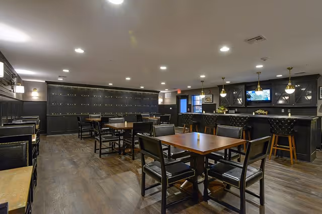 Interior view of a dining area with several wooden tables and black chairs arranged neatly on a dark wooden floor. The room features a bar counter with high black cushioned stools, pendant lights hanging from the ceiling, and a TV mounted on the wall behind the bar. The walls are painted dark with decorative paneling, and there is a door and window visible in the background.