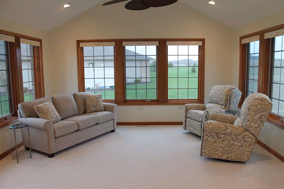 A bright living room with large windows showing a green lawn outside. The room contains a beige sofa with two patterned cushions on the left and two patterned armchairs on the right, each with a small round glass side table next to them. The walls are light-colored and the window frames are wooden.