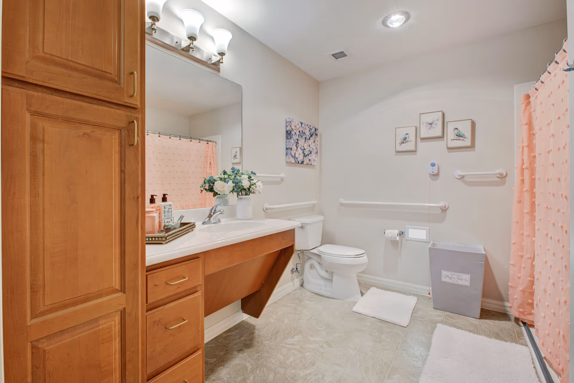 A clean and well-lit bathroom featuring a wooden vanity with a large mirror and three light fixtures above it. The vanity has a white countertop with a vase of flowers and soap dispensers. There is a white toilet next to the vanity, with grab bars installed on the walls for support. The shower area has a pink textured shower curtain. The floor is tiled, and there are white bath mats placed near the toilet and shower. The walls are decorated with framed pictures of birds and flowers. A gray laundry basket is positioned near the shower.