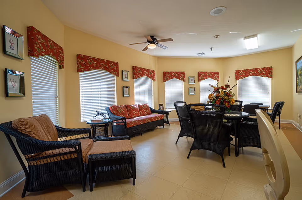 A bright and cozy common area with multiple windows covered by red floral valances. The room features black wicker furniture including a loveseat with red floral cushions, a chair with a matching ottoman, and a round table surrounded by six chairs. A ceiling fan with a light is mounted on the ceiling, and a floral arrangement decorates the center of the table. The walls are painted a soft yellow, and framed artwork is hung on the walls.