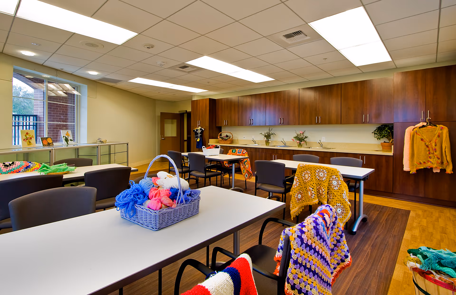 A bright craft room with several tables and chairs, some draped with colorful crocheted blankets. A basket filled with balls of yarn sits on one table. The room has wooden cabinets along one wall with sinks and potted plants, and large windows letting in natural light.