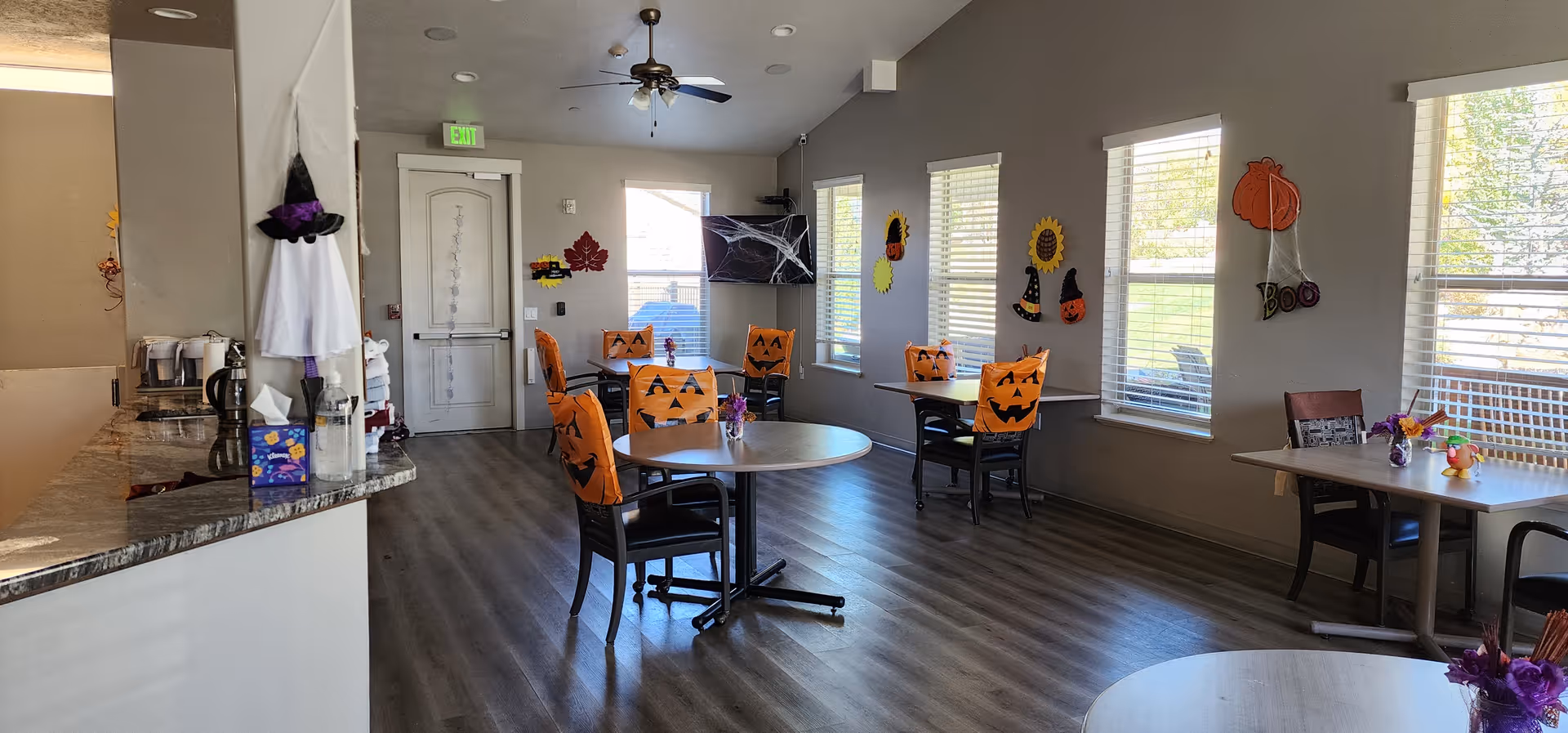 A dining area decorated for Halloween with orange chair covers featuring jack-o'-lantern faces. The room has several tables and chairs, large windows with blinds, and Halloween decorations on the walls including pumpkins, witches' hats, and a 'Boo' sign. There is a counter on the left side with a coffee maker and other items, and a door at the far end of the room.