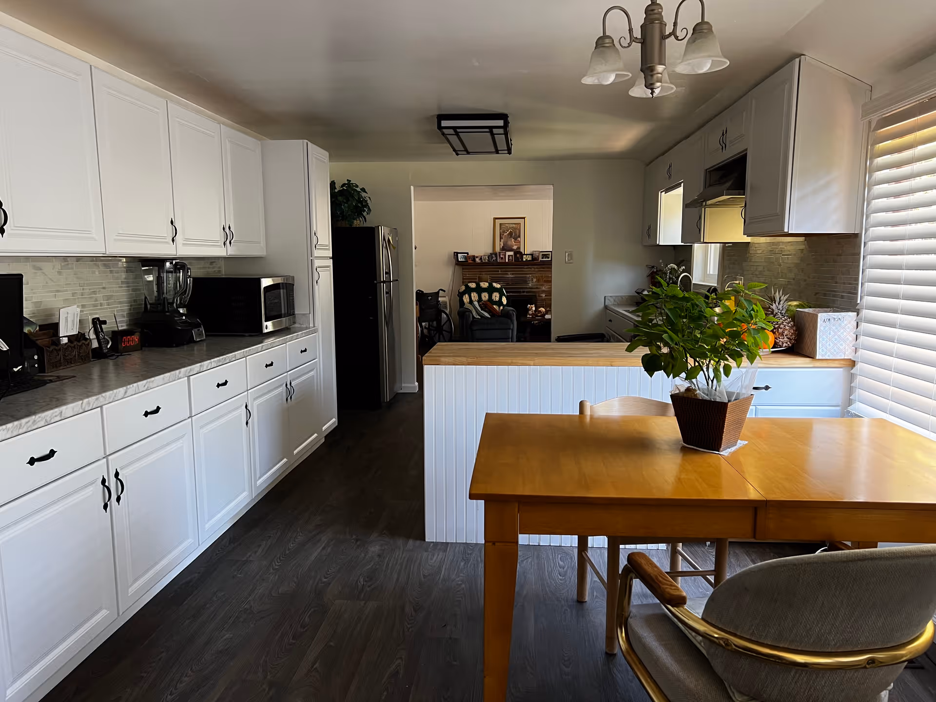 A bright kitchen with white cabinets and a marble countertop. There is a wooden dining table with a potted plant on it near a window with blinds. The kitchen has a refrigerator, microwave, blender, and other small appliances. In the background, there is a view into a living room area with a chair and some framed pictures on a mantel.