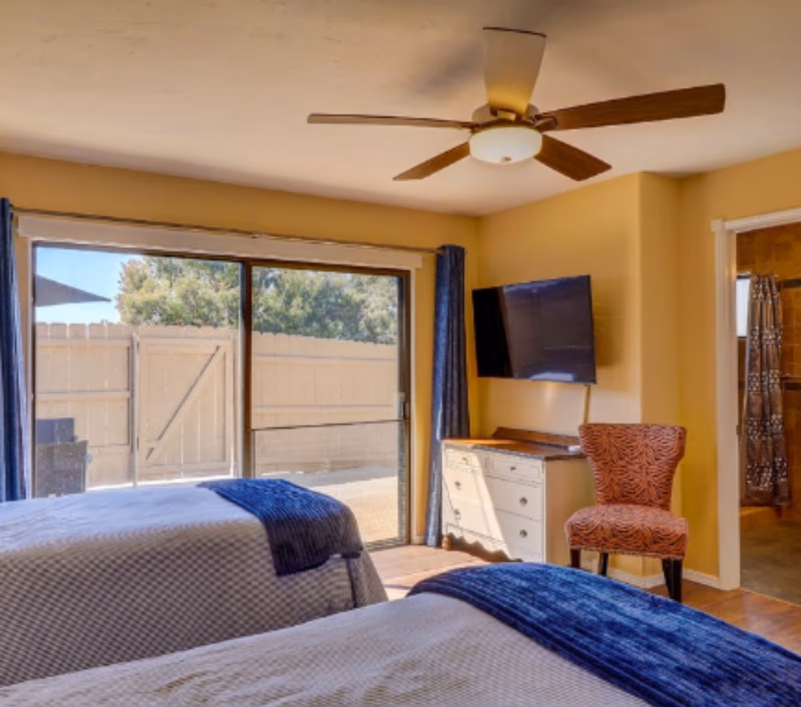 Sunlit bedroom with two beds, a ceiling fan, wall-mounted TV, dresser and sliding glass doors to a fenced patio.