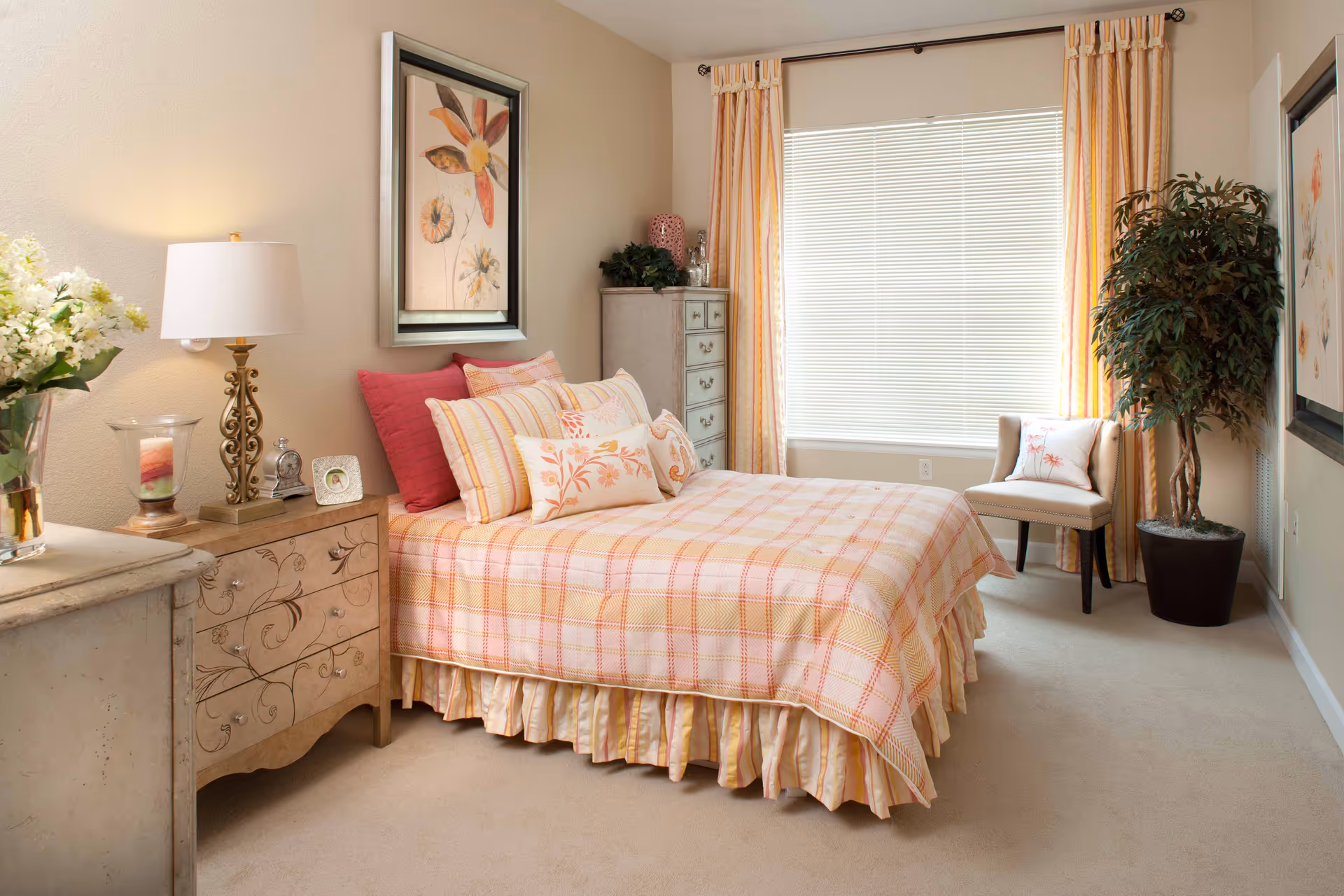 A cozy bedroom with a bed covered in a pink and yellow plaid bedspread and multiple pillows. There is a nightstand with a decorative lamp, a candle, and a small framed photo. A tall dresser stands in the corner next to a window with closed blinds and yellow-striped curtains. A beige upholstered chair with a floral pillow and a large potted plant are placed near the window. The walls are decorated with framed floral artwork.
