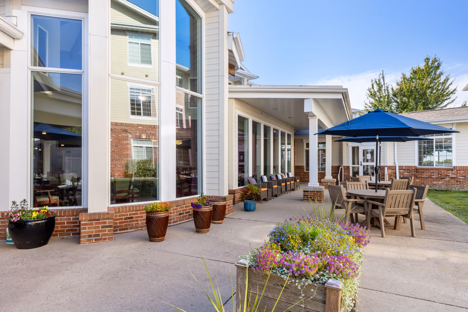 Outdoor patio area at Legacy Pointe with several wooden tables and chairs under blue umbrellas, potted plants and flowers along the walkway, and large windows reflecting the building exterior.
