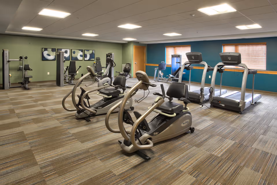 A spacious fitness room with exercise equipment including recumbent bikes, treadmills, and weight machines. The room has green and teal walls, carpeted flooring, and two windows with wooden blinds letting in natural light. There are three abstract paintings on one wall.