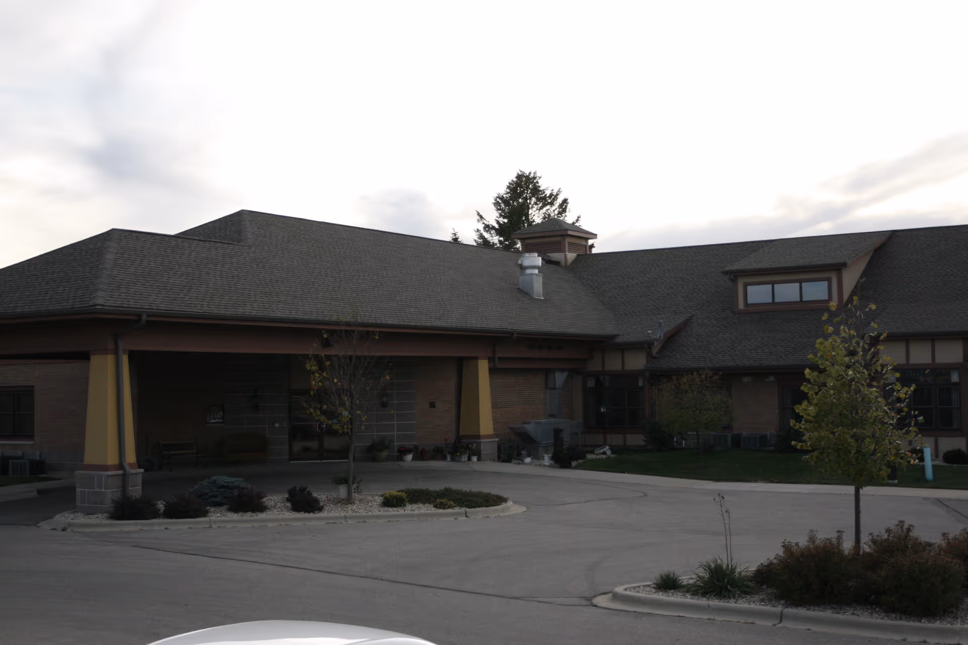 Exterior view of a single-story assisted living facility building with a large covered entrance, surrounded by a paved driveway and landscaped areas with small trees and shrubs under a cloudy sky.