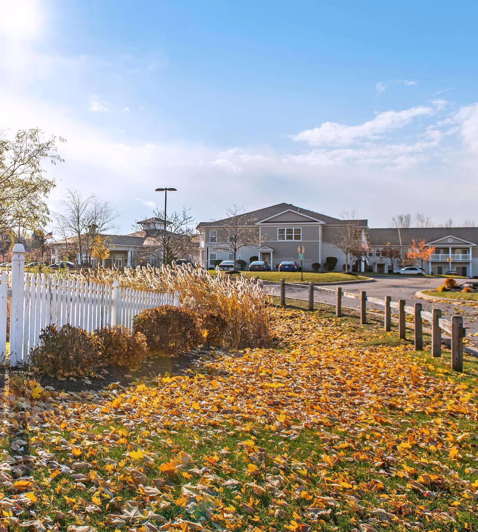 Front view of a senior living building with a white picket fence, autumn leaves on the lawn, and parked cars.