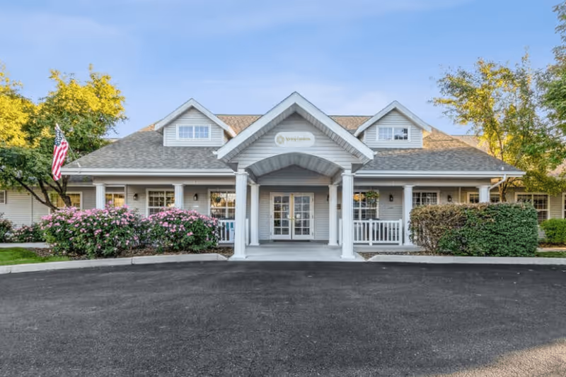 Front exterior view of a single-story senior living facility building with a covered entrance supported by white columns. The building has a gray roof with dormer windows and light gray siding. There are bushes with pink flowers and green shrubs on either side of the entrance, and an American flag is visible on the left side. The sky is clear and blue.