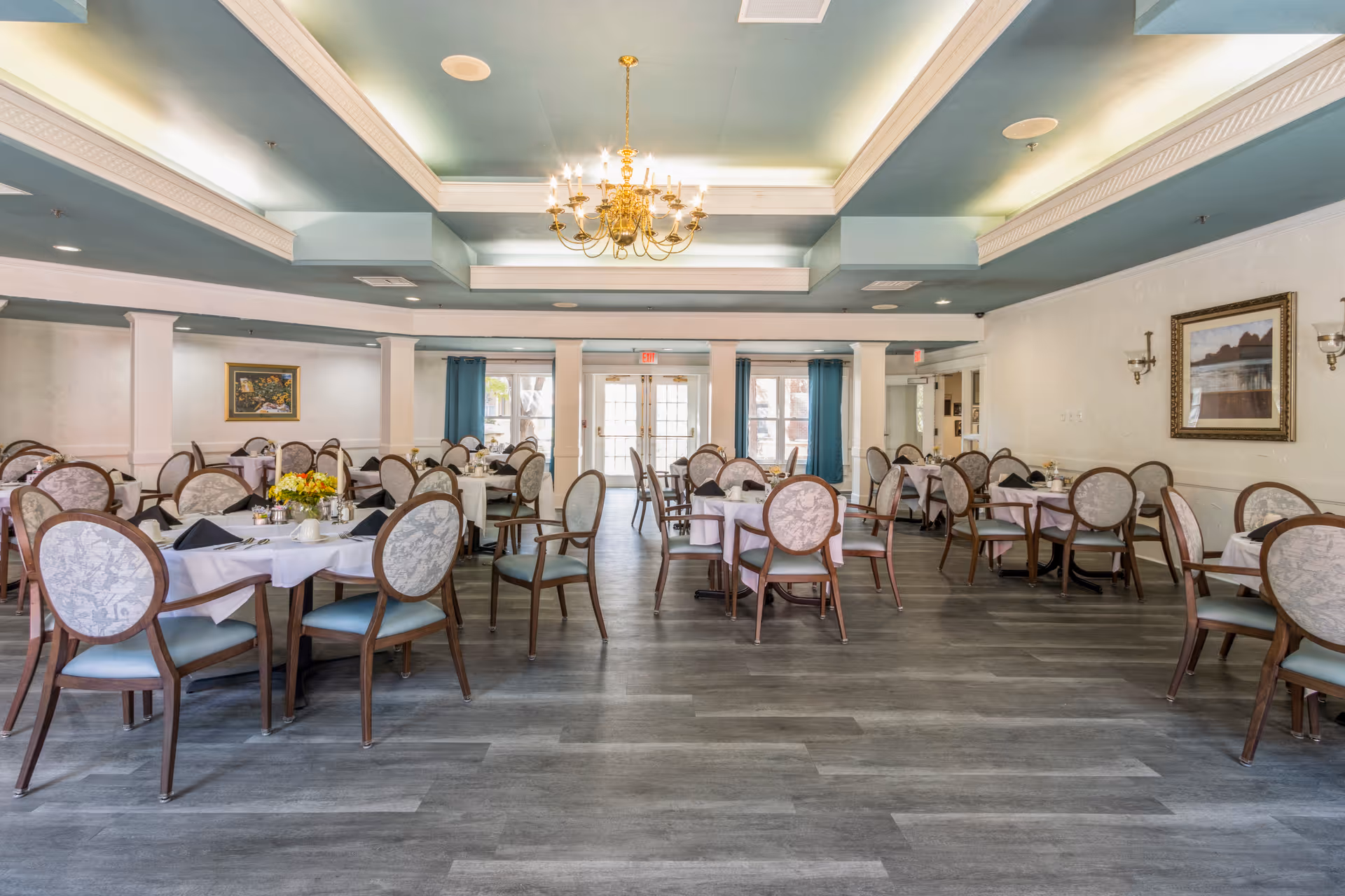 A spacious dining room with multiple round tables covered with white tablecloths and set with cups, plates, and napkins. The chairs have wooden frames and upholstered backs and seats. The room features a decorative ceiling with a chandelier, light blue ceiling accents, and large windows with blue curtains allowing natural light to enter. There are framed paintings on the walls and sconces providing additional lighting.