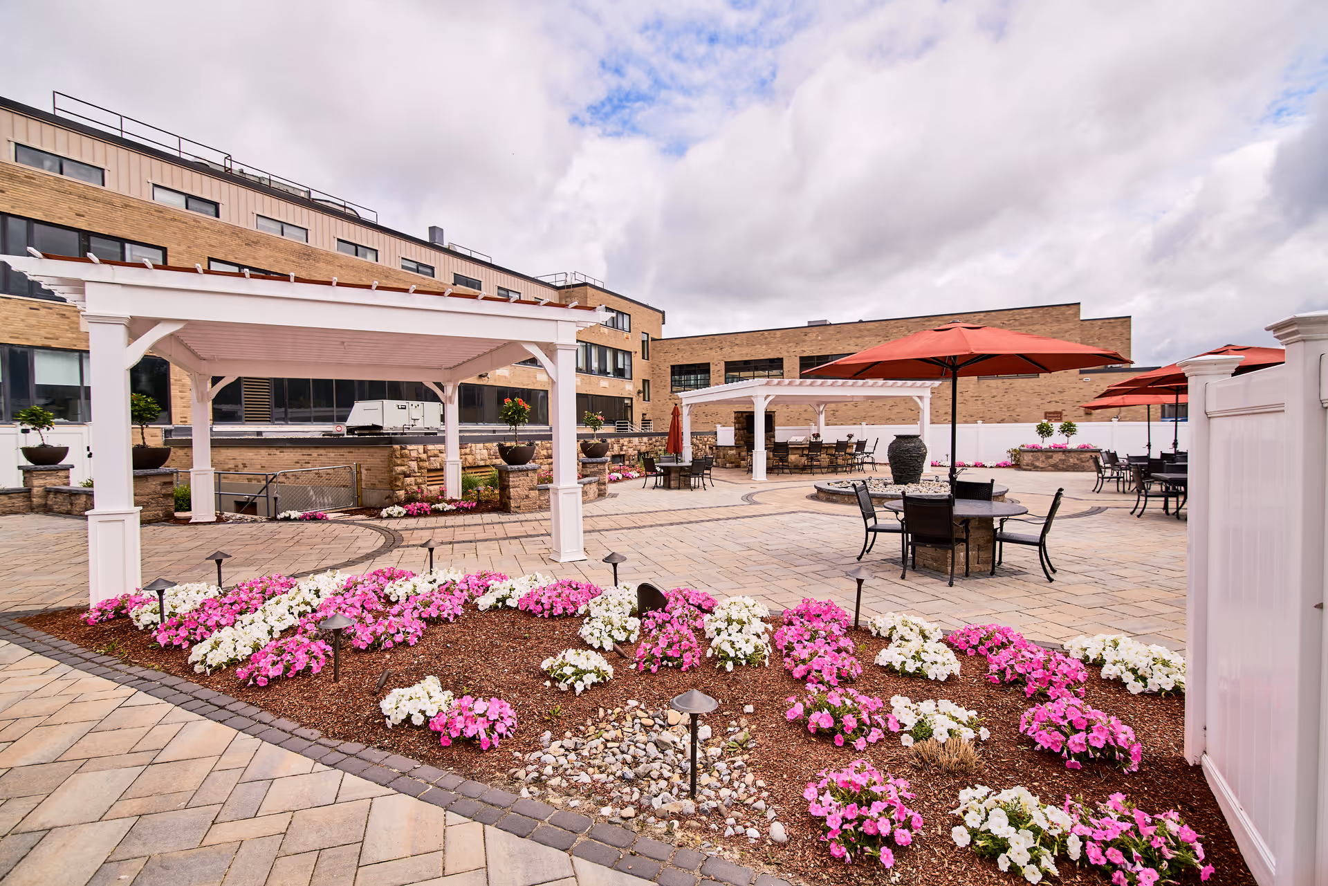 Outdoor patio area at CareOne at Concord featuring paved walkways, white pergolas, round tables with red umbrellas, black chairs, and flower beds with pink and white flowers under a cloudy sky.
