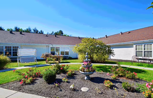Outdoor garden area at Kessler Estates Senior Living featuring a circular flower bed with a tiered fountain in the center, surrounded by green grass, bushes, and flowers. There is a white swing bench with blue and white striped cushions on the left side and several chairs along the building walls. The building has white siding and a brown roof under a clear blue sky.