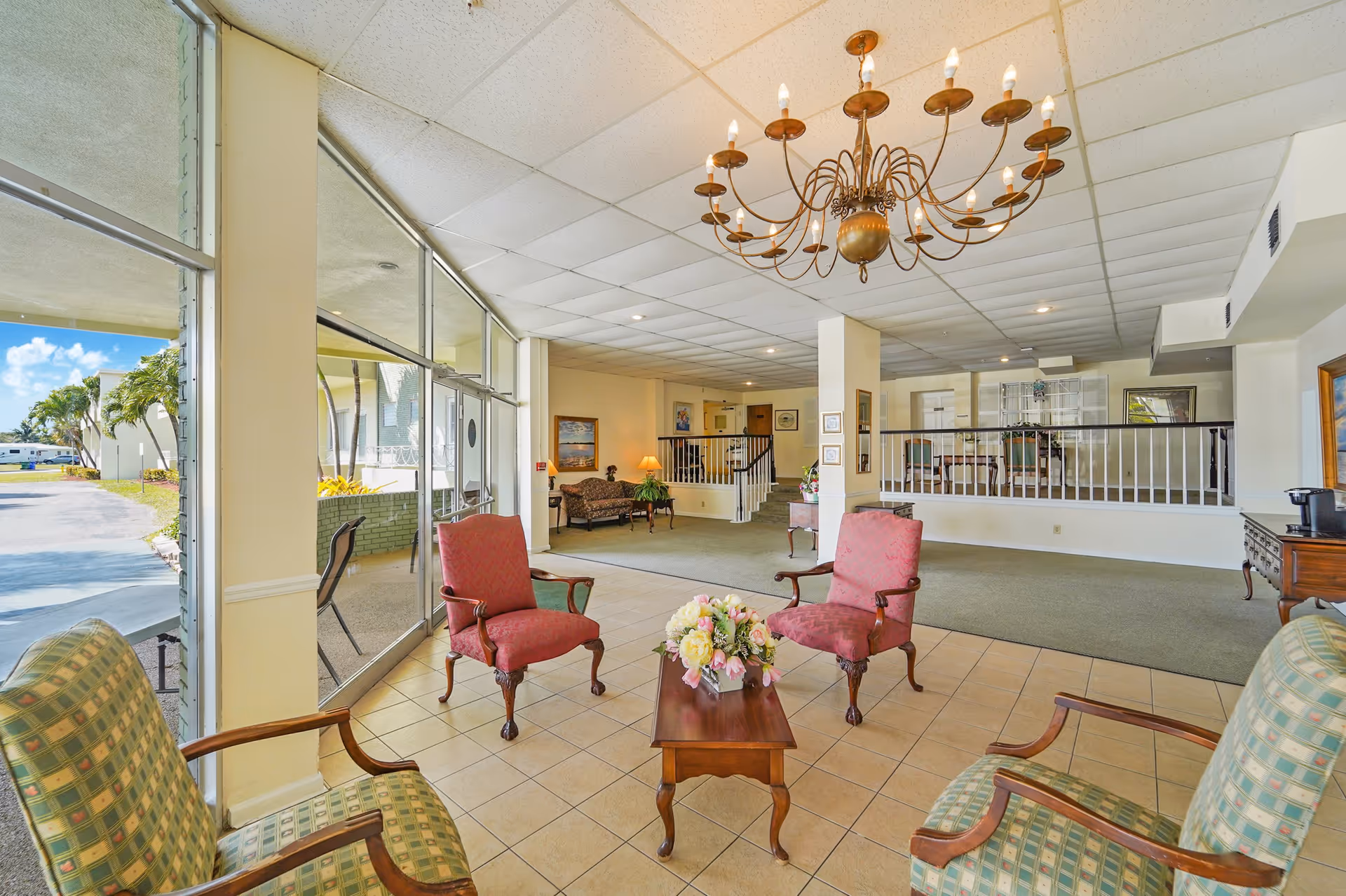 A spacious and well-lit assisted living residence common area with large windows letting in natural light. The room features a tiled floor section with four upholstered chairs arranged around a wooden coffee table with a floral centerpiece. In the background, there is a carpeted area with additional seating, a staircase, and decorative lamps. A large chandelier hangs from the ceiling.