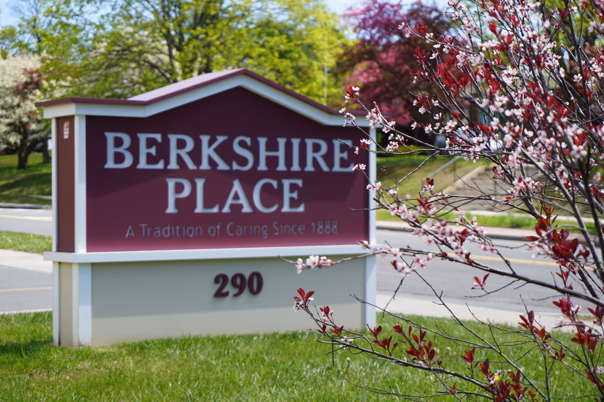 Outdoor sign for Berkshire Place with the tagline 'A Tradition of Caring Since 1888' and the number 290, surrounded by green grass and flowering trees.