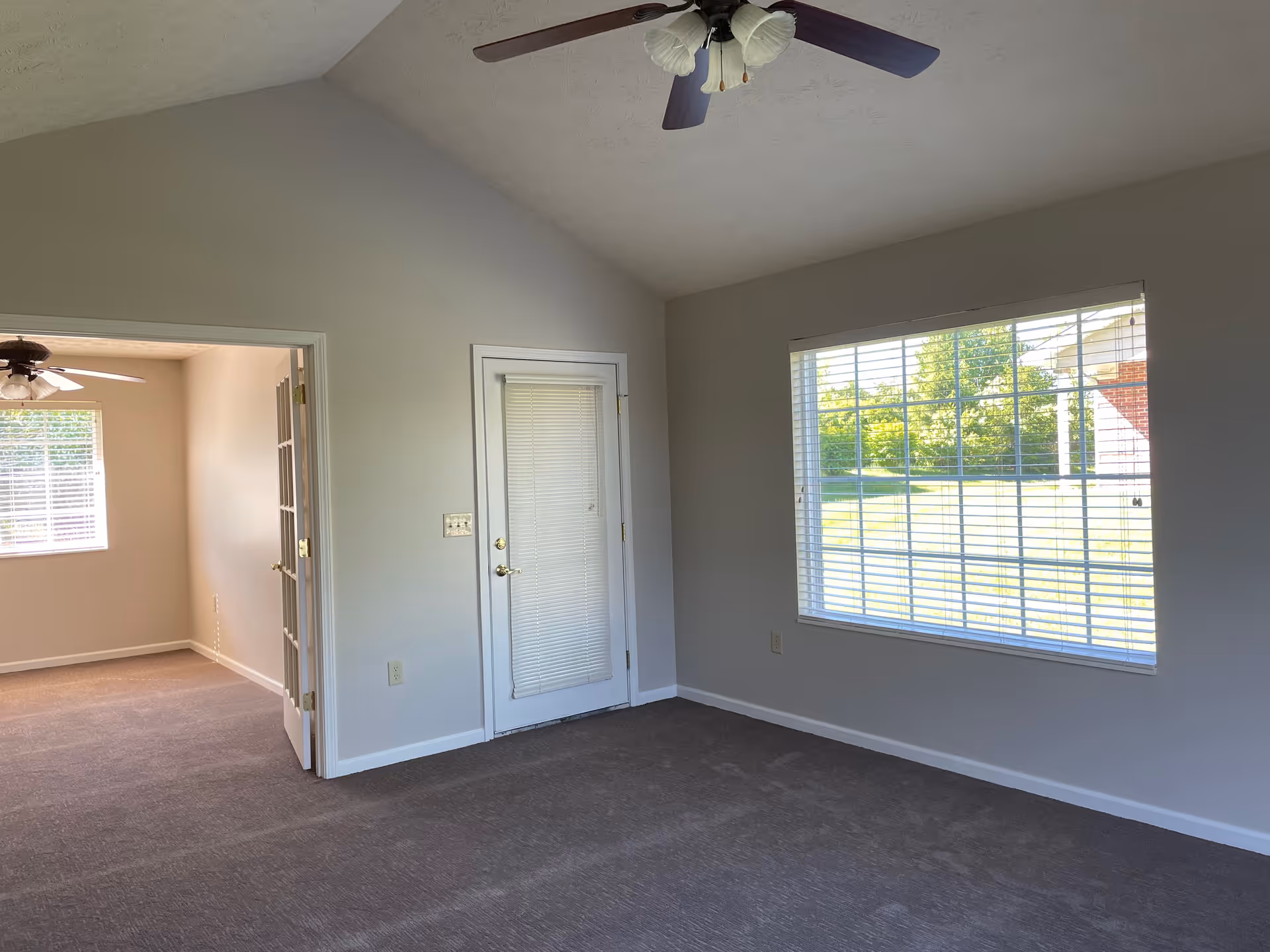 Empty carpeted living room with a ceiling fan, a door with built-in blinds, and a large window overlooking a lawn.