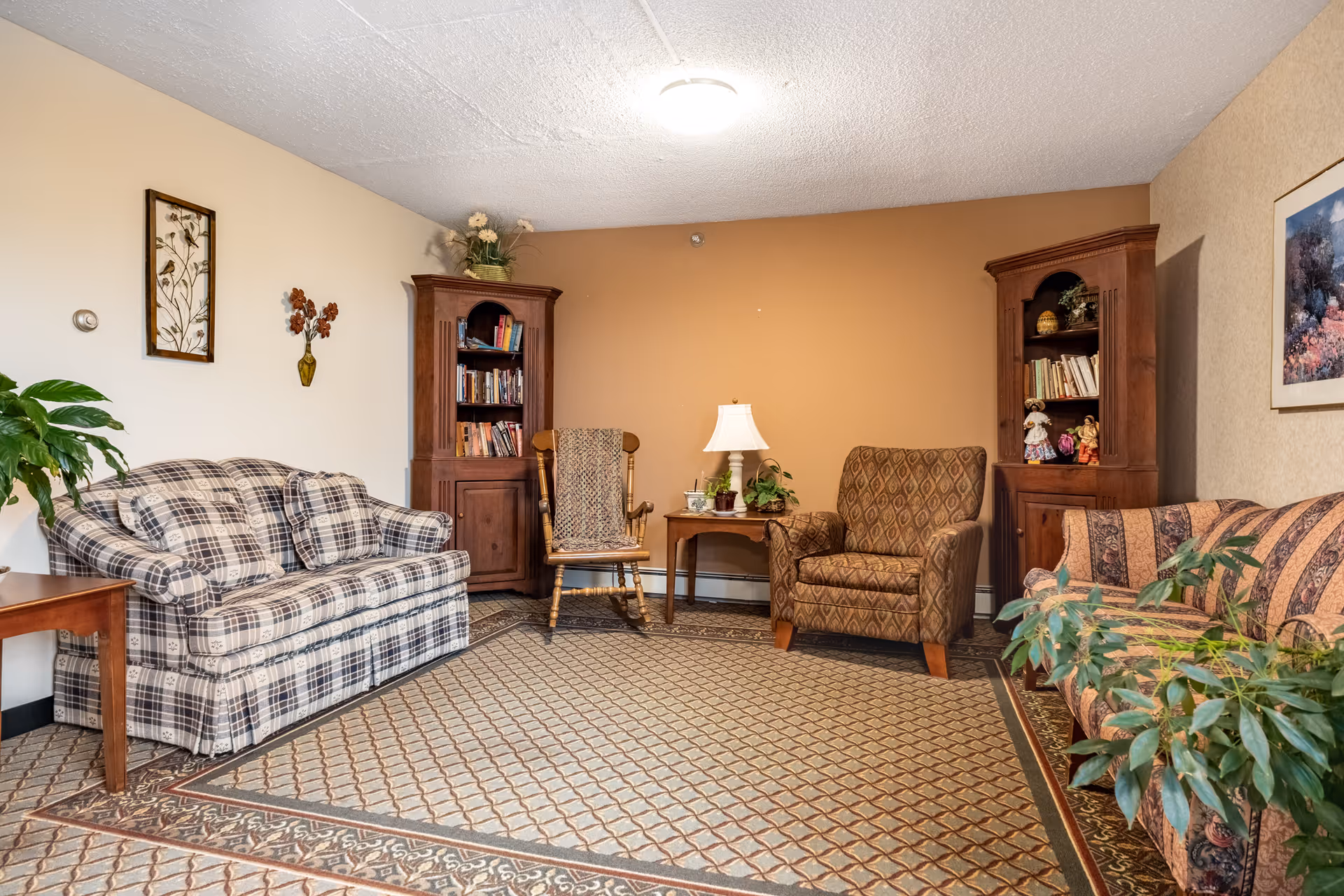A cozy living room in a senior living facility featuring a plaid sofa, a patterned armchair, a wooden rocking chair, two wooden bookcases filled with books and decorative items, a small wooden side table with a lamp and plants, wall art, and a large patterned area rug covering the floor.