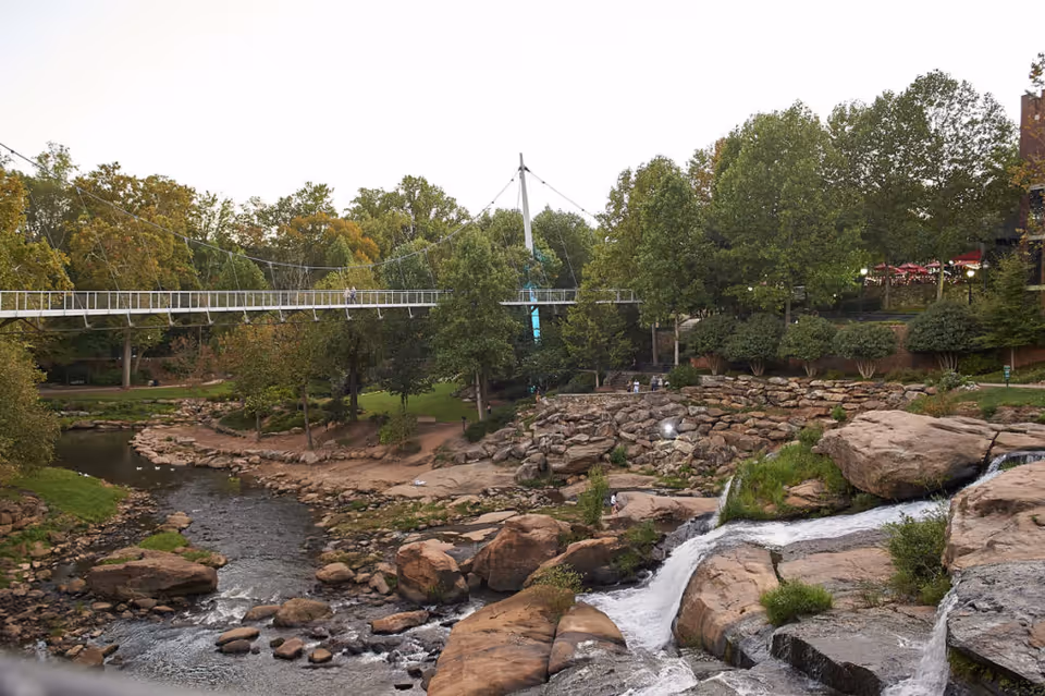 A scenic outdoor park area featuring a flowing river with rocks and small waterfalls, surrounded by green trees and bushes. A suspension pedestrian bridge spans across the river, and there are people walking and sitting in the park. The setting appears peaceful and natural.