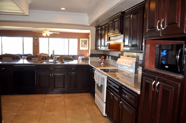A spacious kitchen with dark wooden cabinets, a white stove, a microwave, and a granite countertop. The kitchen opens up to a living area with large windows letting in natural light.