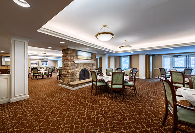 Spacious dining room with multiple tables and chairs arranged neatly, featuring a central stone fireplace, carpeted floor with a patterned design, large windows with curtains allowing natural light, and ceiling lights providing warm illumination.