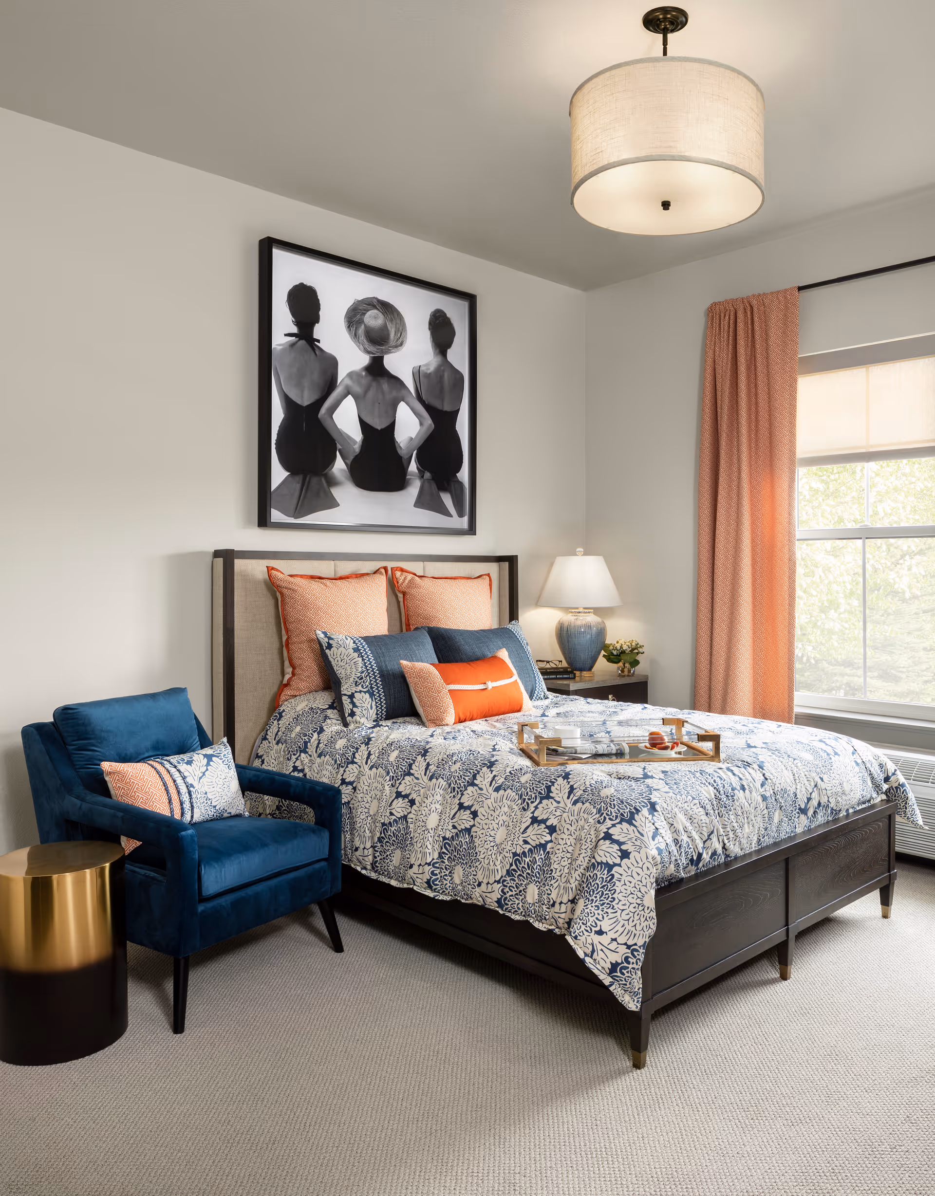 Well-decorated bedroom with a bed dressed in blue-and-white patterned bedding and coral pillows, a blue armchair, framed black-and-white artwork above the headboard, and a window with coral curtains.