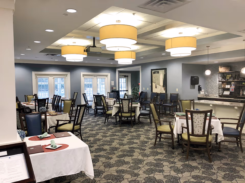 A dining room in a senior living facility with multiple tables covered with white tablecloths, each set with placemats, napkins, and menus. The room has patterned carpet, blue and green cushioned chairs, large round ceiling lights, and windows with white blinds. There is a counter area with shelves and decorative items on the right side of the room.