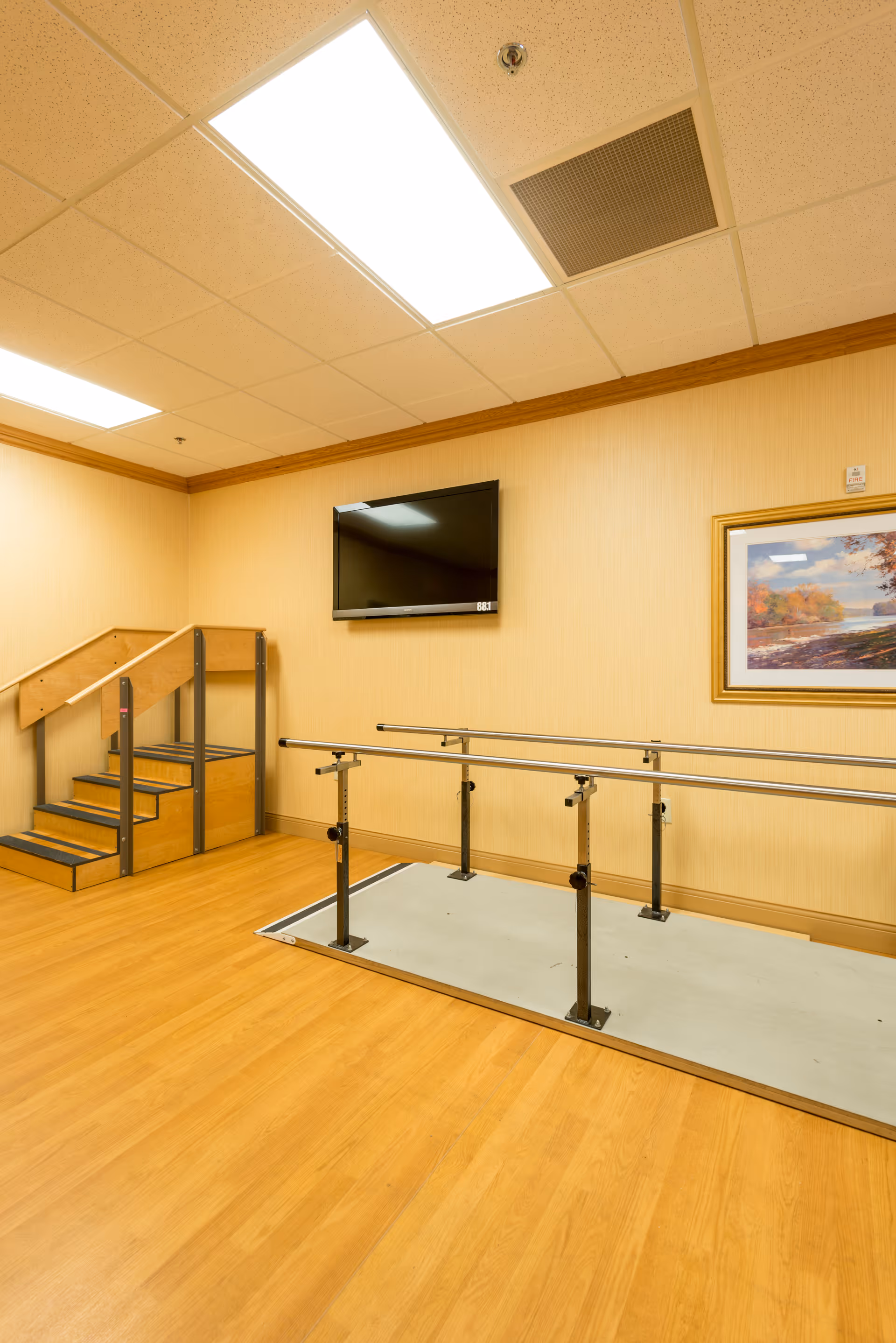 Physical therapy room with parallel walking bars, a small practice staircase, wall-mounted TV, and framed art.
