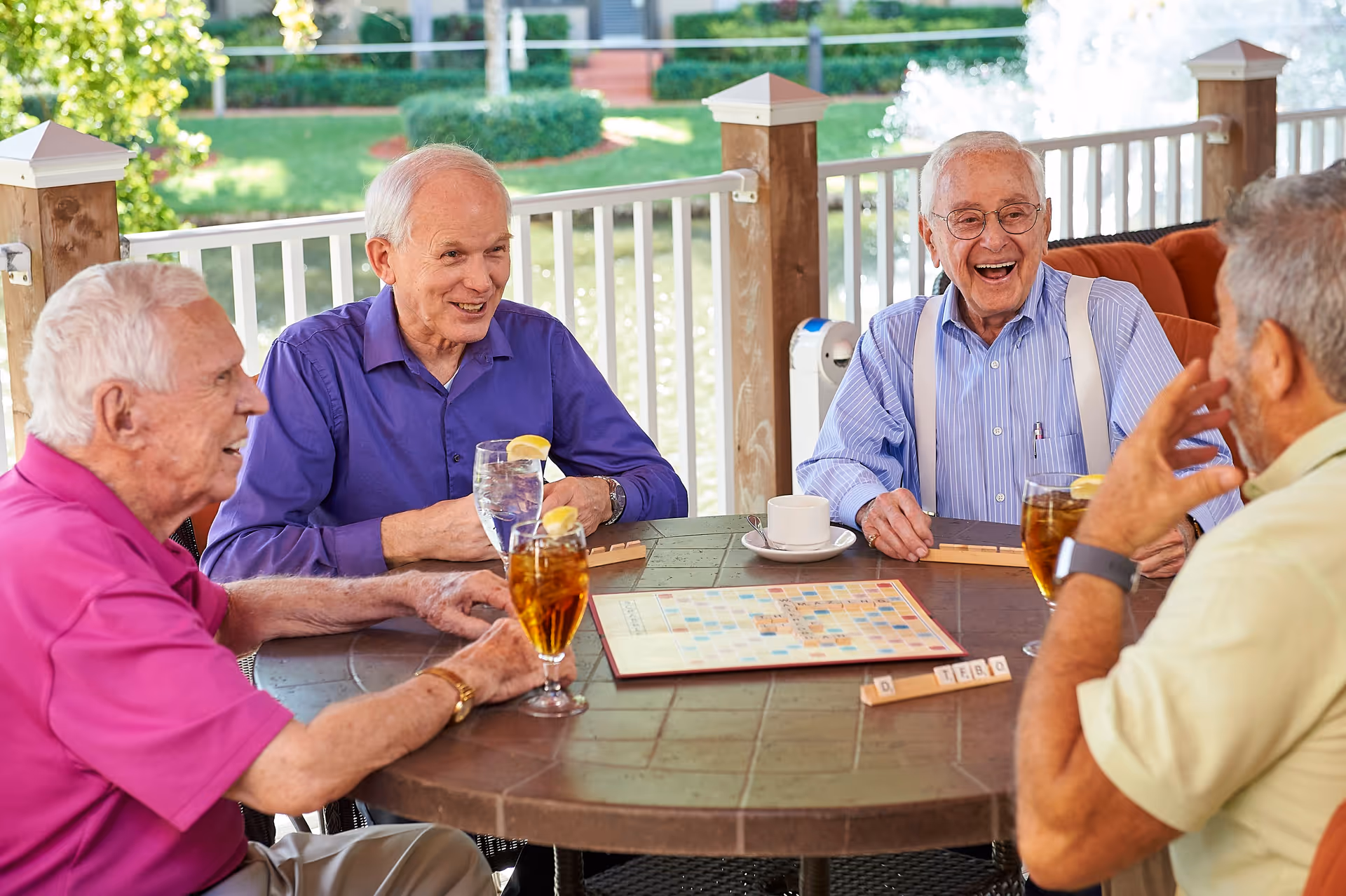 Four elderly men sitting around a round outdoor table playing a word game similar to Scrabble. They are smiling and engaged in conversation. The table has drinks with lemon slices and a coffee cup. The setting is a covered patio with a white railing and greenery in the background.