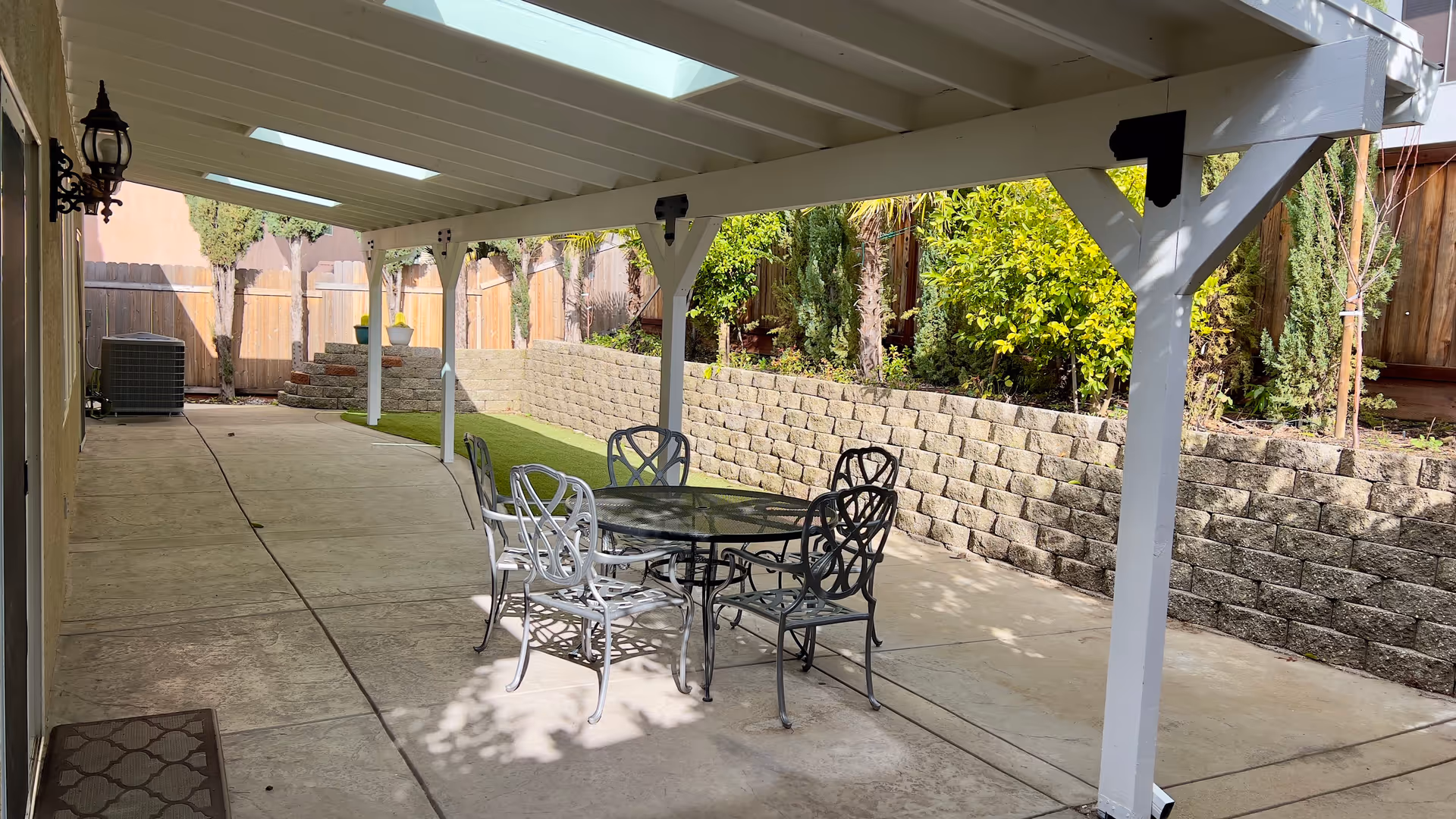 Covered backyard patio with a glass-top table and metal chairs under a white pergola beside a stone retaining wall and wooden fence.