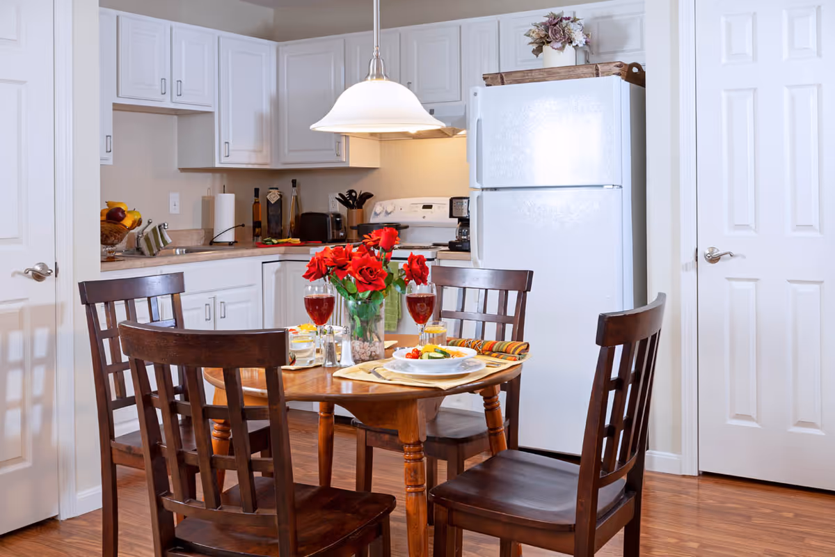 A cozy kitchen and dining area with white cabinets, a white refrigerator, and a stove. A wooden dining table with four matching chairs is set with plates of food, glasses of red wine, and a vase of red flowers in the center. The floor is wooden, and there are two white doors on either side of the kitchen.