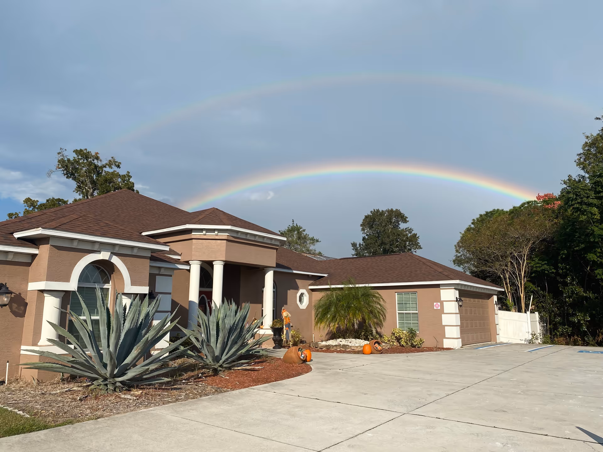 Exterior view of Cobblestone Manor Assisted Living Facility showing a single-story building with a brown roof and beige walls. The driveway and parking area are visible in the foreground, with some landscaping including large agave plants and a small palm tree. There are pumpkins and fall decorations near the entrance. A double rainbow arcs across the sky above the building.