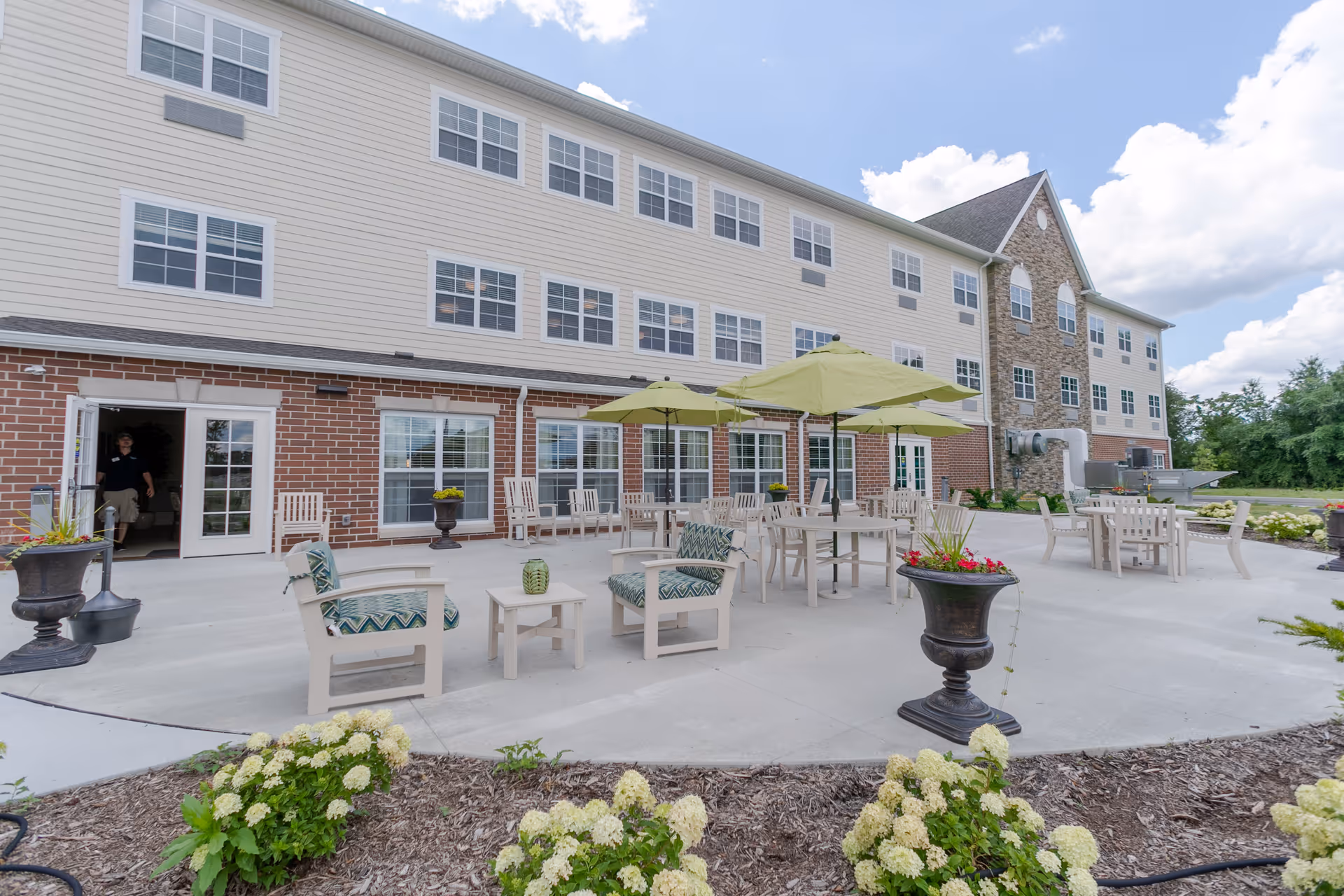 Outdoor patio with tables, chairs, umbrellas and potted plants in front of a multi-story senior living building.