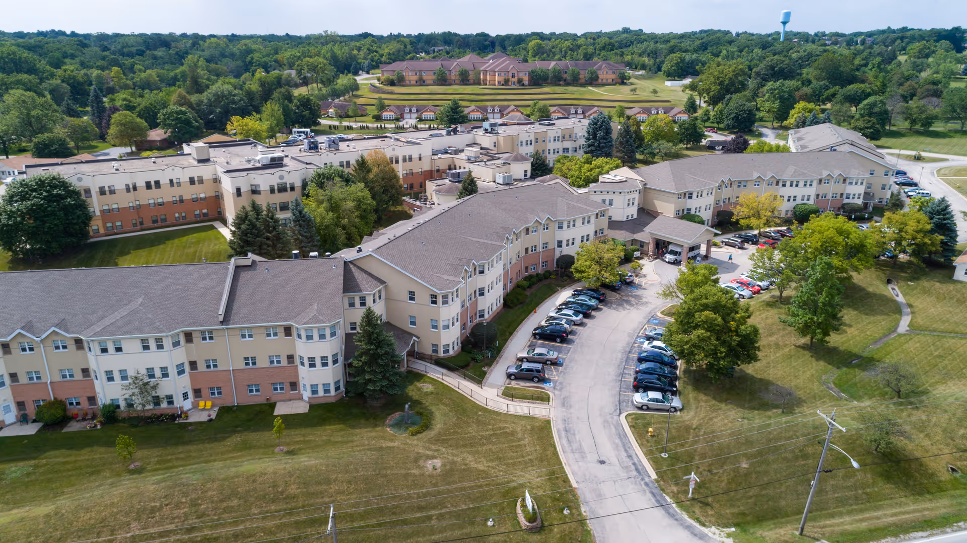 Aerial view of Franciscan Village senior living facility showing multiple connected beige and light brown buildings with a curved driveway and parked cars. The facility is surrounded by green lawns, trees, and a wooded area in the background under a clear sky.
