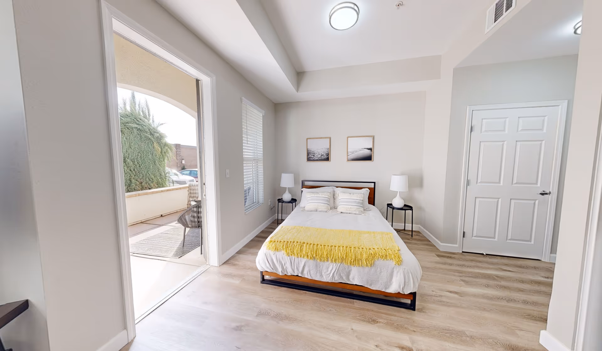 A bright bedroom with a double bed centered against the back wall, adorned with white bedding and a yellow throw blanket. Two matching bedside tables with white lamps flank the bed. Above the bed are two framed pictures. To the left, a door opens to an outdoor patio area with chairs and greenery visible. The room has light-colored wood flooring and neutral-colored walls.