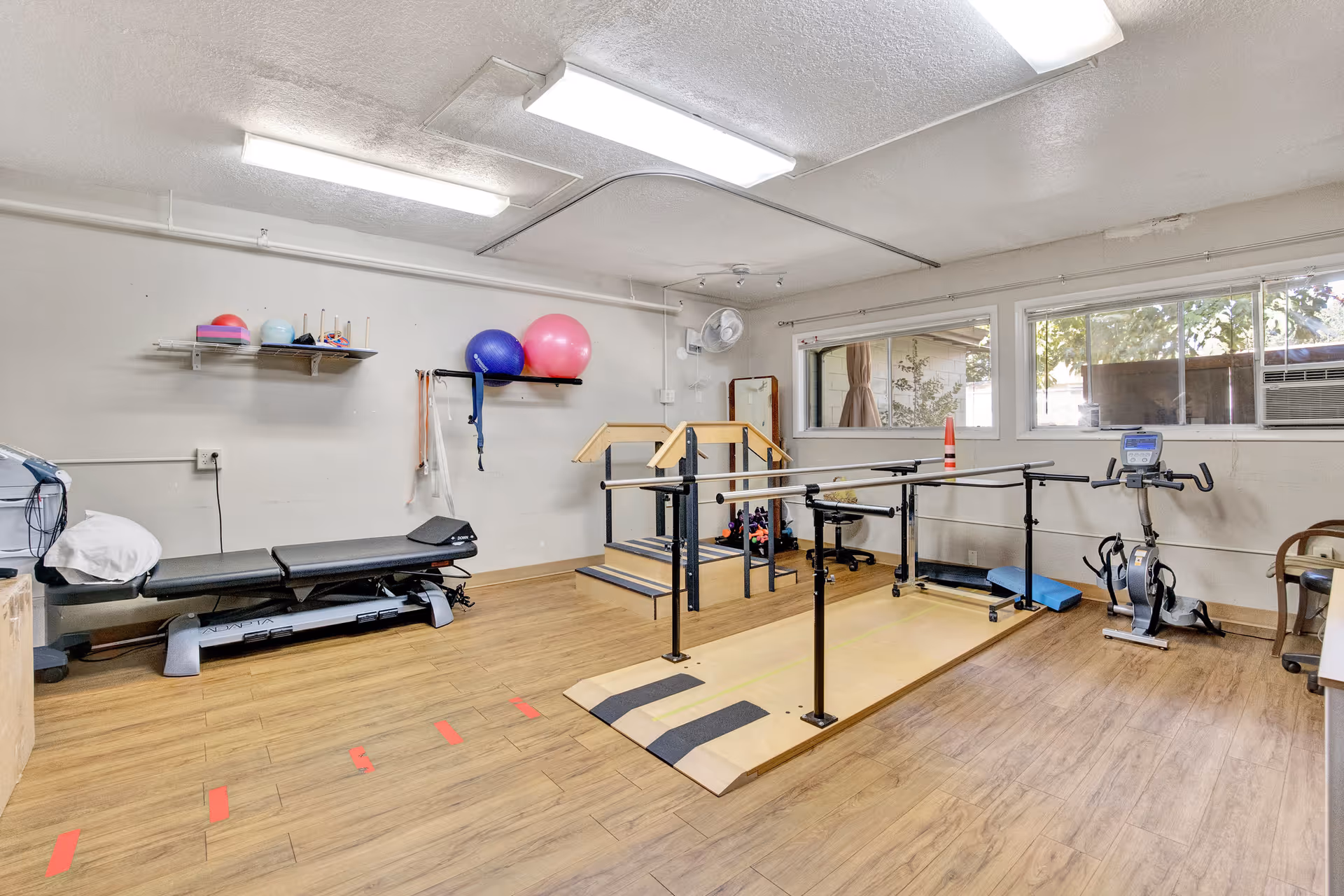 Rehabilitation therapy room with parallel bars, treatment table, exercise bike, and colorful exercise balls.
