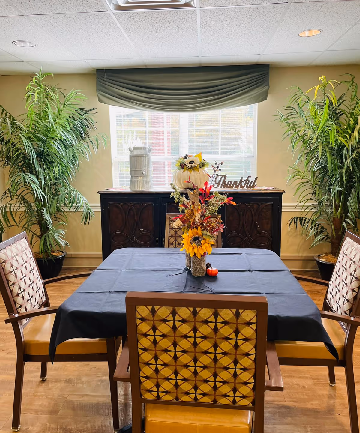 A dining area with a square table covered by a black tablecloth, surrounded by four chairs with patterned backs and yellow seats. On the table is a floral centerpiece with sunflowers and autumn-themed decorations. Behind the table is a dark wooden sideboard with a white pumpkin decoration and a sign that says 'Thankful'. Two large green potted plants flank the sideboard, and a window with blinds and a green valance is in the background.