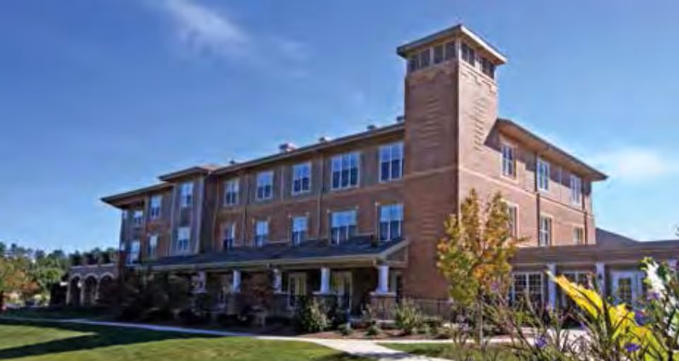 Exterior view of a three-story brick building with multiple windows and a tower-like structure on one corner, surrounded by landscaped greenery and a clear blue sky.