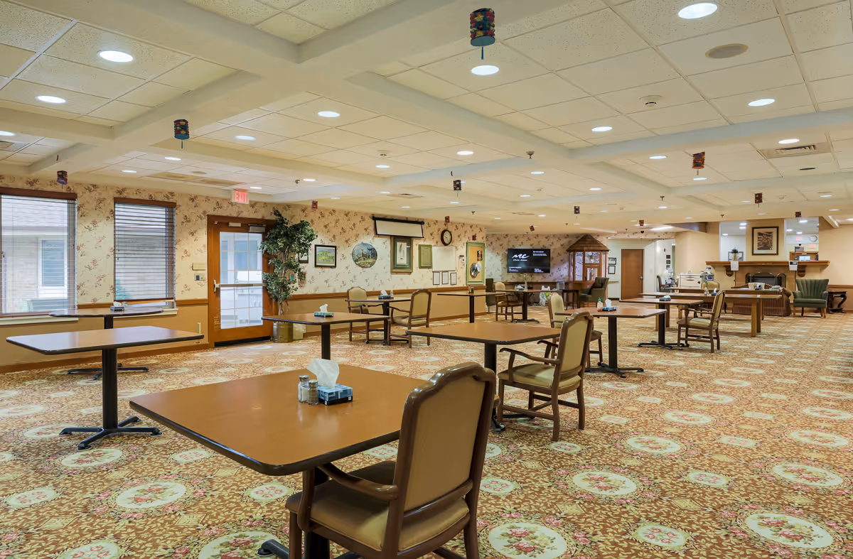 Spacious dining room with multiple square wooden tables and cushioned chairs arranged neatly. The room has patterned carpet flooring, floral wallpaper, and a ceiling with recessed lighting. There are windows with blinds on one side and a door with glass panels. The far wall features framed pictures, a clock, and a large TV screen. The room is well-lit and decorated with small hanging ornaments from the ceiling.
