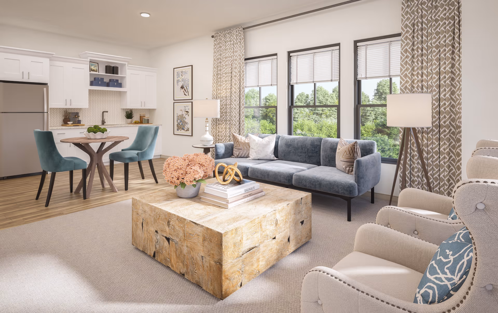 A bright and modern living room and kitchen area with large windows showing green trees outside. The living room features a blue sofa with decorative pillows, two beige armchairs with blue patterned cushions, a wooden coffee table with books and a flower arrangement, and two lamps with white shades. The kitchen area has white cabinets, a stainless steel refrigerator, a small round wooden dining table with two teal upholstered chairs, and a backsplash with a herringbone pattern.