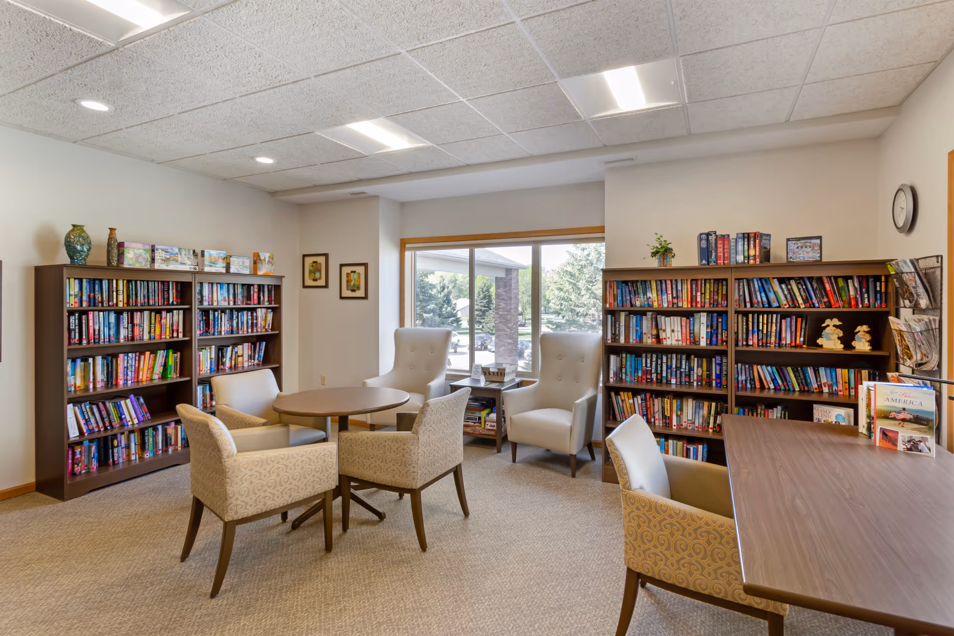 A cozy senior living library room with two large bookshelves filled with books, a round table surrounded by four upholstered chairs, two high-back armchairs near a large window, and a rectangular table with chairs. The room has beige walls, carpeted floor, and ceiling lights.