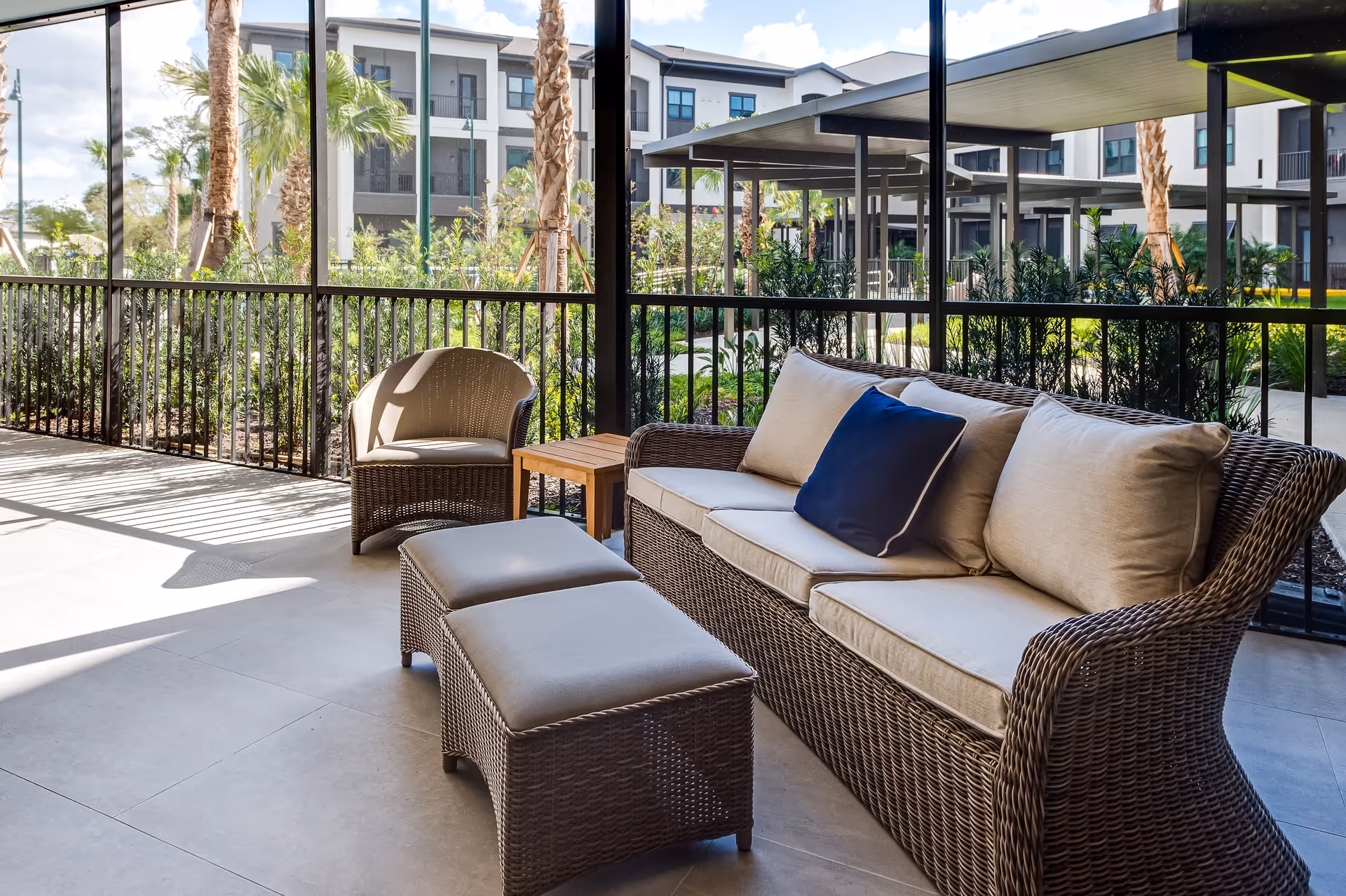 A covered outdoor patio area with wicker furniture including a sofa with beige cushions and a blue pillow, two ottomans, and a chair. The patio overlooks a landscaped garden with palm trees and a multi-story residential building in the background.