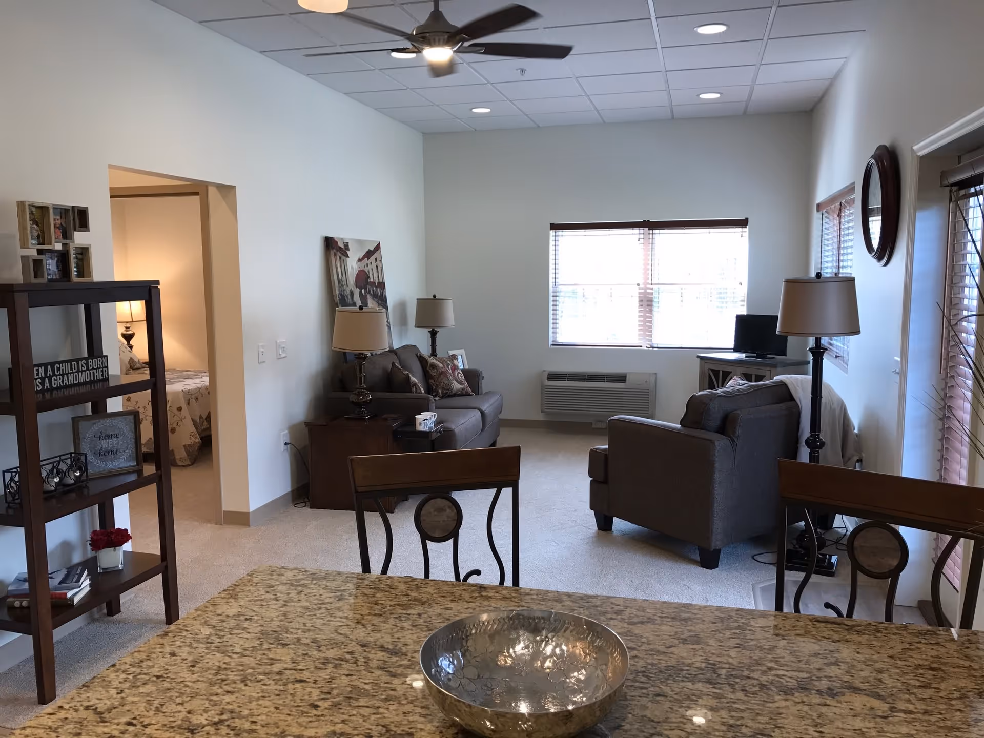 Interior view of a senior living facility apartment showing a living room with two gray sofas, a small TV on a stand, two lamps, a ceiling fan, and a window with blinds. In the foreground, there is a granite countertop with a decorative bowl. To the left, there is a wooden shelving unit with framed pictures and decor, and an open doorway leading to a bedroom with a bed and a lamp.