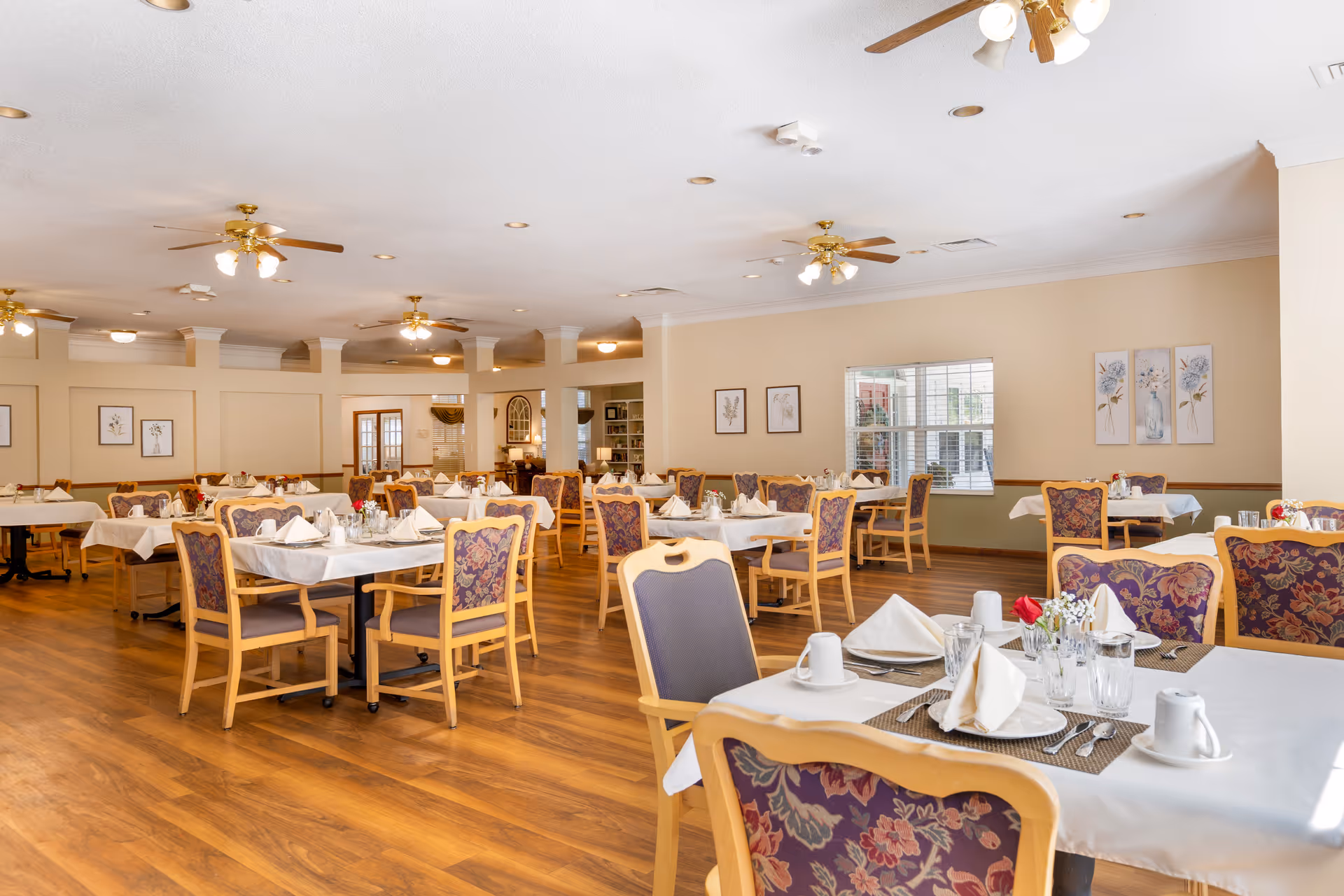 Spacious dining room with multiple set tables and wooden chairs, ceiling fans, and floral artwork on the walls.